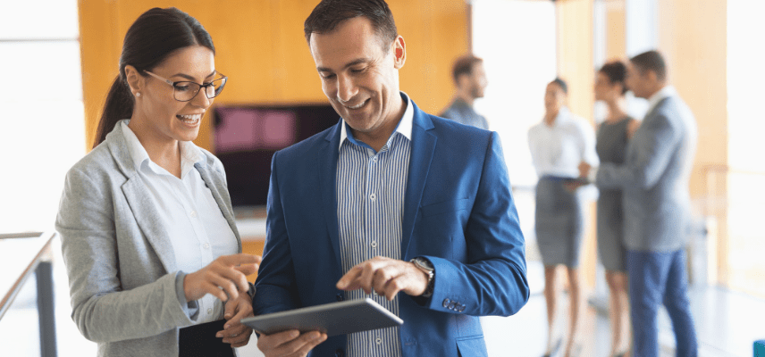 A man and woman point to the screen of a tablet device that he's holding. Coworkers are talking in the background.