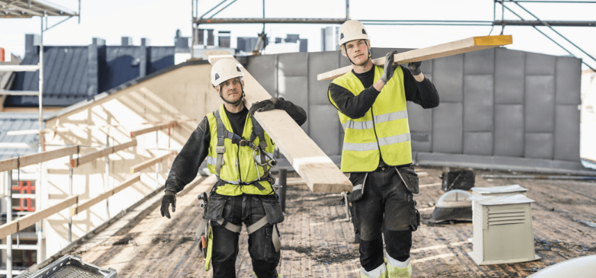 Two construction workers in hard hats carrying lumber at a construction site